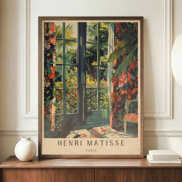 May include: Framed Henri Matisse exhibition poster featuring vibrant tropical scene with French doors opening to lush garden filled with palm fronds, colorful flowers in greens, oranges, and corals. Poster displays on wooden sideboard beside white ceramic vase and stacked books in elegant room with cream walls and white molding.
