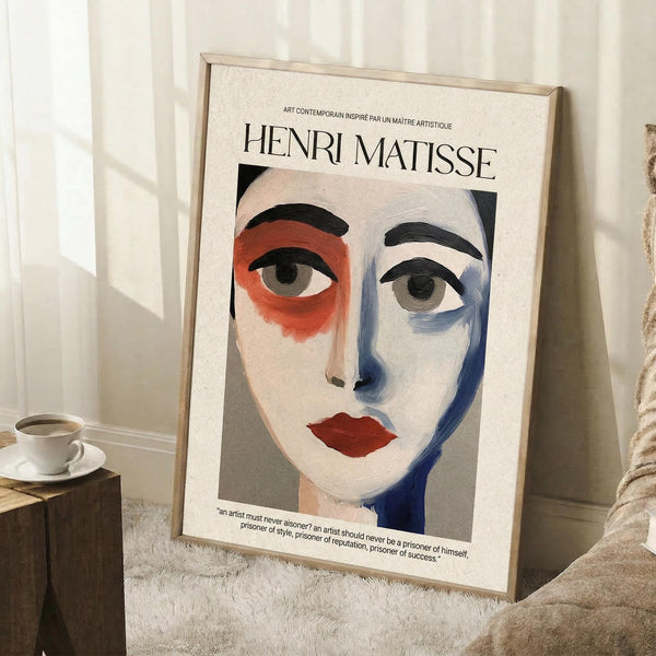 May include: Framed art print featuring an abstract portrait with bold brushstrokes in red, blue, and white tones, showing a stylized face with prominent eyes and red lips. The poster has "HENRI MATISSE" text at the top and includes a quote at the bottom. Displayed in a light wood frame leaning against white walls on a textured cream rug, with a coffee cup on books nearby and soft natural lighting creating shadows.