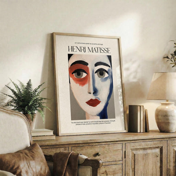 May include: Framed Henri Matisse-inspired portrait poster displayed on rustic wooden console table. Abstract face features red and blue color blocking around eyes, red lips, black hair against cream background. Light wood frame, surrounded by fern plant, leather chair, ceramic vases and books in neutral-toned living room setting.