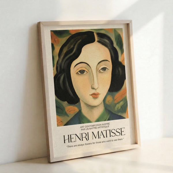 A framed art print featuring a stylized portrait of a woman with short dark wavy hair and pale skin, painted in warm earth tones with green and orange floral elements in the background. The artwork has a modernist style with simplified features and bold colors. Below the image is text reading "HENRI MATISSE" and a quote about flowers. The print is displayed in a light wood frame leaning against a white wall.