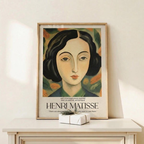 May include: Framed poster print featuring stylized portrait of woman with black bob haircut and coral lips against colorful floral background in muted greens and oranges. Text reads "Henri Matisse" below artwork. Natural wood frame sits on white wooden surface next to small wrapped gift with dried sprig. Soft lighting creates gentle shadows on cream wall background.