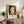 May include: Framed art print featuring a portrait of a woman with dark wavy hair and green-orange floral background, displayed on rustic wooden console table alongside fern plant, ceramic vases, and books in neutral-toned living room setting with beige chair and pillows