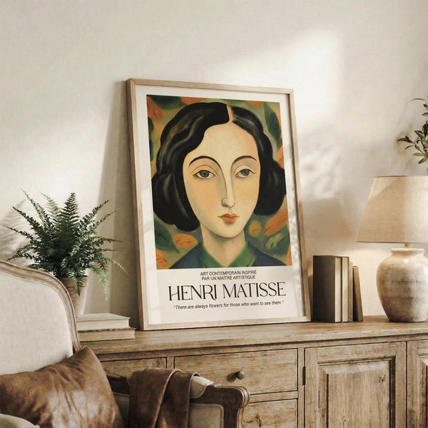 May include: Framed art print featuring a portrait of a woman with dark wavy hair and green-orange floral background, displayed on rustic wooden console table alongside fern plant, ceramic vases, and books in neutral-toned living room setting with beige chair and pillows