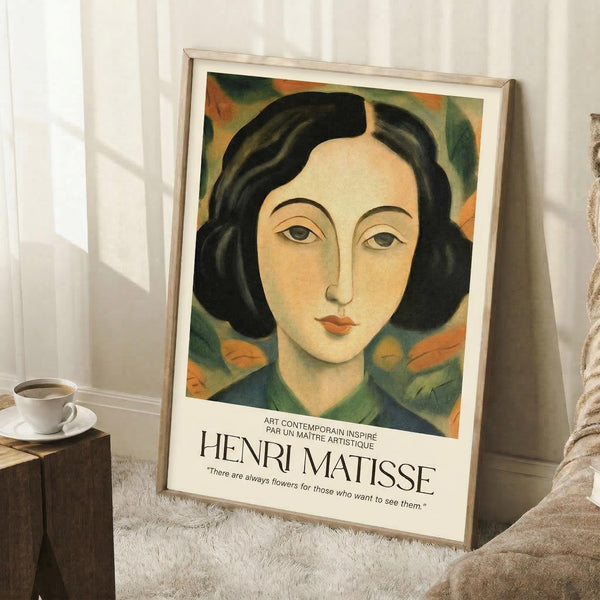 May include: Framed portrait poster in light wood frame leaning against white wall on textured white rug. Artwork shows woman with dark bob haircut, pale complexion, and colorful floral background in greens and oranges. Poster has white border with Henri Matisse text and quote below image. Coffee cup on wooden block visible to left, beige throw blanket on right side of scene.