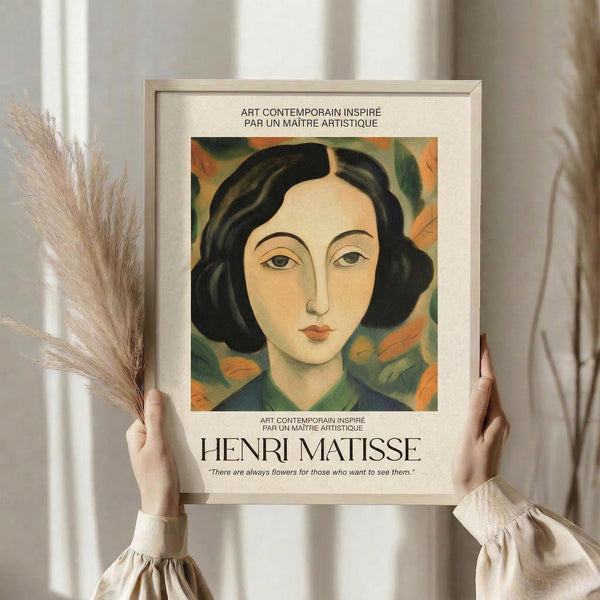 May include: Person holding framed art print featuring portrait of woman with black wavy hair and coral lips against green and orange floral background, styled as vintage Matisse-inspired poster with French text and artist quote, displayed in light wooden frame in bright interior setting with dried pampas grass