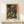 May include: Framed art print of a bearded man's portrait in vibrant colors including orange, green, and blue brushstrokes, displayed on a white wooden shelf next to a small wrapped gift box with twine and greenery sprig against a cream wall.