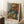 May include: A framed art print featuring a colorful portrait of a bearded man in Fauve style with bold oranges, greens, and blues, displayed on a white fluffy rug against white walls with natural window light, alongside a coffee cup on a wooden surface and cream-colored fabric.