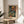 May include: Framed art print of a colorful self-portrait showing a bald man with mustache and beard in vibrant oranges, blues, and greens. The poster sits on a round wooden side table next to a beige upholstered chair, with plant leaves visible in soft natural lighting creating shadows on a white wall.