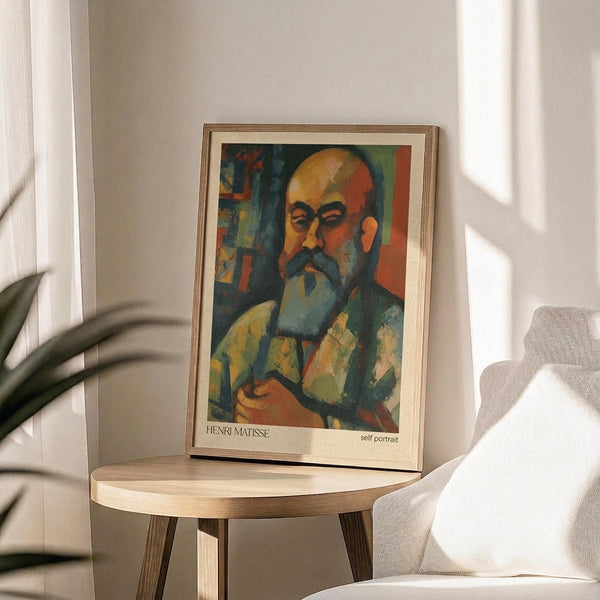 May include: Framed art print of a colorful self-portrait showing a bald man with mustache and beard in vibrant oranges, blues, and greens. The poster sits on a round wooden side table next to a beige upholstered chair, with plant leaves visible in soft natural lighting creating shadows on a white wall.