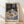 May include: Framed art print of a still life painting showing a round table with blue patterned tablecloth, white teapot with blue floral design, bowls of colorful oranges and fruits, black mug, and decorative plates. Blue patterned wallpaper visible in background. Natural wood frame displayed on light wooden floor against white wall with soft shadows.