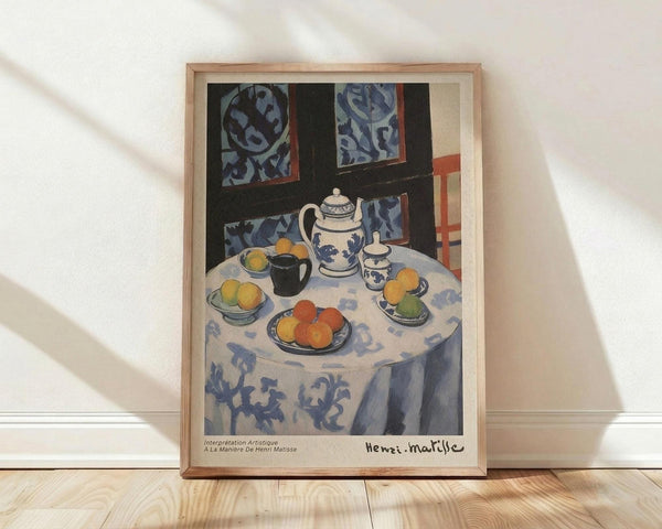 May include: Framed art print of a still life painting showing a round table with blue patterned tablecloth, white teapot with blue floral design, bowls of colorful oranges and fruits, black mug, and decorative plates. Blue patterned wallpaper visible in background. Natural wood frame displayed on light wooden floor against white wall with soft shadows.