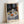 May include: Framed art print leaning against white wall showing Henri Matisse still life painting with blue and white patterned tablecloth, ceramic teapot, colorful oranges and fruit on plates, dark window frames with blue patterned curtains in background. Light wooden frame with white mat and artist signature visible at bottom.