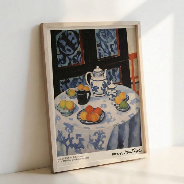 May include: Framed art print leaning against white wall showing Henri Matisse still life painting with blue and white patterned tablecloth, ceramic teapot, colorful oranges and fruit on plates, dark window frames with blue patterned curtains in background. Light wooden frame with white mat and artist signature visible at bottom.