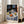 May include: Framed Henri Matisse still life print leaning against white wall next to beige armchair. Artwork shows round table with blue patterned tablecloth, white teapot with blue designs, colorful fruit bowls with oranges and lemons, black mug, and decorative wallpaper background. Natural wood frame, bright room with large windows casting sunlight on hardwood floors.