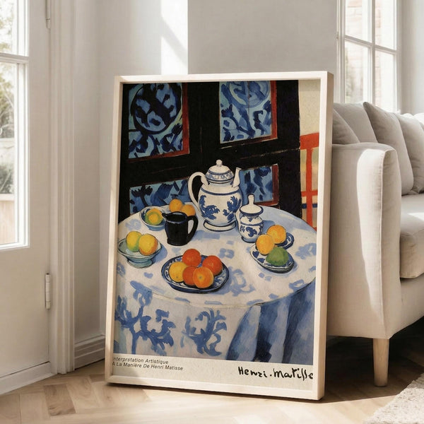 May include: Framed Henri Matisse still life print leaning against white wall next to beige armchair. Artwork shows round table with blue patterned tablecloth, white teapot with blue designs, colorful fruit bowls with oranges and lemons, black mug, and decorative wallpaper background. Natural wood frame, bright room with large windows casting sunlight on hardwood floors.