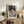 May include: Framed Henri Matisse still life print displayed on rustic wooden sideboard in cozy living room setting. Artwork features blue and white patterned tablecloth with tea service, oranges, and decorative elements in Matisse's distinctive Post-Impressionist style. Scene includes fern plant, ceramic vase, lamp, books, and cream upholstered chair with brown throw pillows against neutral wall.