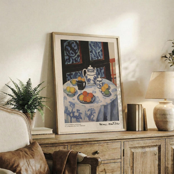 May include: Framed Henri Matisse still life print displayed on rustic wooden sideboard in cozy living room setting. Artwork features blue and white patterned tablecloth with tea service, oranges, and decorative elements in Matisse's distinctive Post-Impressionist style. Scene includes fern plant, ceramic vase, lamp, books, and cream upholstered chair with brown throw pillows against neutral wall.