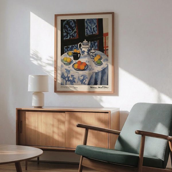 May include: Framed Henri Matisse still life print showing a round table with white and blue patterned tablecloth, tea set, fruit bowls, and decorative window panels in background. Displayed above wooden sideboard with table lamp in modern living room setting with sage green mid-century chair and natural lighting casting shadows on white wall.