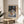 May include: Framed Henri Matisse poster print displayed on round wooden side table next to beige upholstered chair. The artwork shows a colorful still life scene with a white teapot, fruit on plates, and decorative patterned wallpaper in blues and oranges. Natural sunlight creates soft shadows across the neutral-toned room setting with visible plant leaves in corner.
