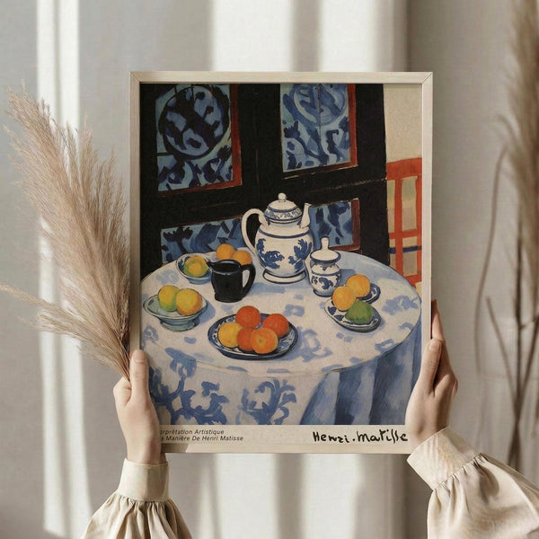 May include: Person holding framed art print showing colorful still life painting with blue and white patterned round table, teapot, cups, bowls of oranges and lemons, decorative background with floral motifs in blues and oranges, displayed against white curtained window with dried pampas grass nearby