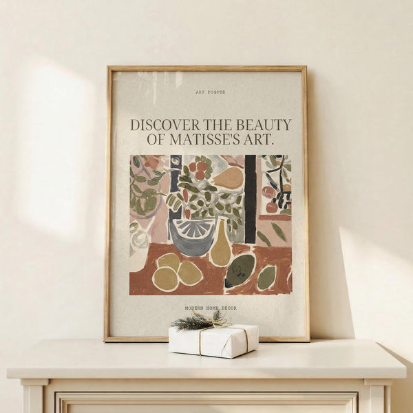 May include: Framed art poster featuring Matisse-inspired still life with plants, vases, and fruits in earthy tones of green, terracotta, and cream. Text reads "Discover the Beauty of Matisse's Art" above colorful botanical illustration. Natural wood frame displayed on light wooden surface with small wrapped gift box beside it against cream wall with soft shadows.