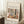 May include: Framed art poster leaning against white wall corner on textured rug. Features colorful still life artwork with plants, fruits, and decorative objects in earthy tones of green, brown, and cream. Text reads "Discover the Beauty of Matisse's Art" above the illustration. Natural light creates shadows on wall. Coffee cup on wooden block and beige fabric visible nearby.
