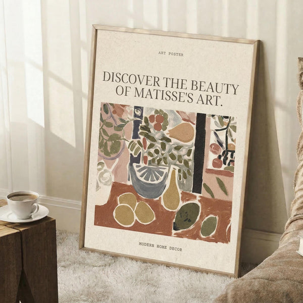 May include: Framed art poster leaning against white wall corner on textured rug. Features colorful still life artwork with plants, fruits, and decorative objects in earthy tones of green, brown, and cream. Text reads "Discover the Beauty of Matisse's Art" above the illustration. Natural light creates shadows on wall. Coffee cup on wooden block and beige fabric visible nearby.