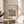 May include: Framed art poster featuring Henri Matisse-style still life with colorful botanical elements, fruits, and vessels in earthy tones of terracotta, green, and cream. Natural wood frame sits on round wooden side table next to beige upholstered chair. Soft natural lighting creates gentle shadows from nearby plant leaves. Minimalist neutral room setting with white curtains and walls.