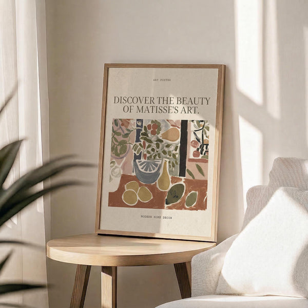 May include: Framed art poster featuring Henri Matisse-style still life with colorful botanical elements, fruits, and vessels in earthy tones of terracotta, green, and cream. Natural wood frame sits on round wooden side table next to beige upholstered chair. Soft natural lighting creates gentle shadows from nearby plant leaves. Minimalist neutral room setting with white curtains and walls.