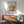 May include: Matisse-inspired art poster with colorful still life featuring fruits, plants and geometric shapes in earthy tones, displayed in thin wooden frame above mid-century modern walnut credenza with white ceramic lamp, next to sage green upholstered armchair with wooden arms in sunlit room with white walls