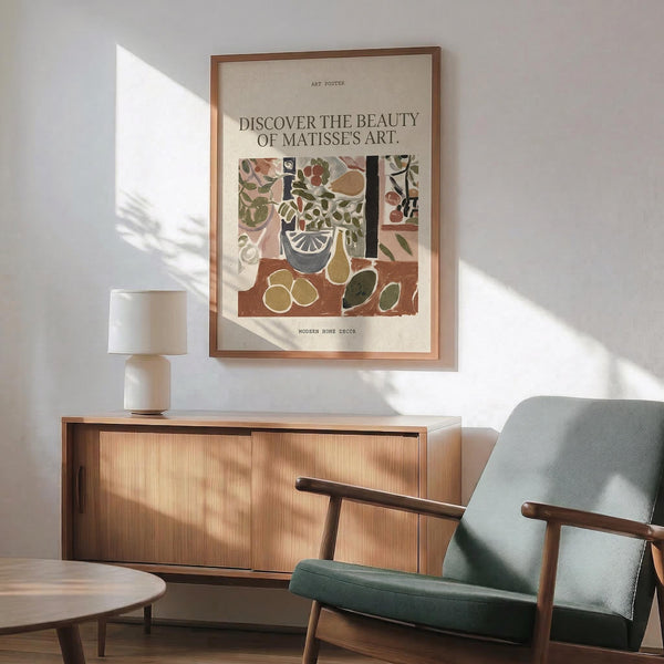 May include: Matisse-inspired art poster with colorful still life featuring fruits, plants and geometric shapes in earthy tones, displayed in thin wooden frame above mid-century modern walnut credenza with white ceramic lamp, next to sage green upholstered armchair with wooden arms in sunlit room with white walls