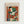May include: Framed art poster featuring a colorful abstract portrait in Matisse's distinctive style, showing a figure wearing a striped hat rendered in bold blocks of green, orange, red, white and black. The minimalist composition uses geometric shapes and vibrant colors against a warm orange background. Displayed in a light wood frame on a white wall.
