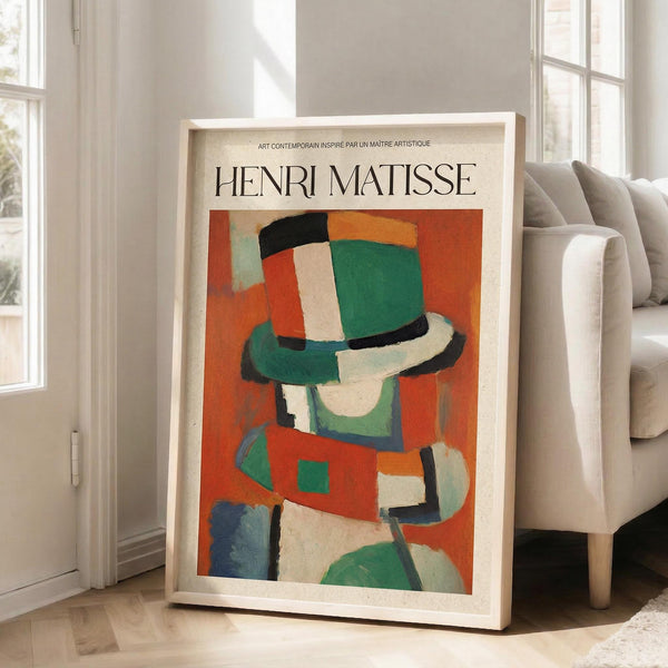 May include: Framed Henri Matisse art print leaning against white wall beside beige sectional sofa in bright living room. Abstract figure wearing colorful hat rendered in bold geometric shapes - vibrant oranges, greens, blues, and cream tones. Light wood frame, hardwood floors, large windows with natural sunlight streaming in.