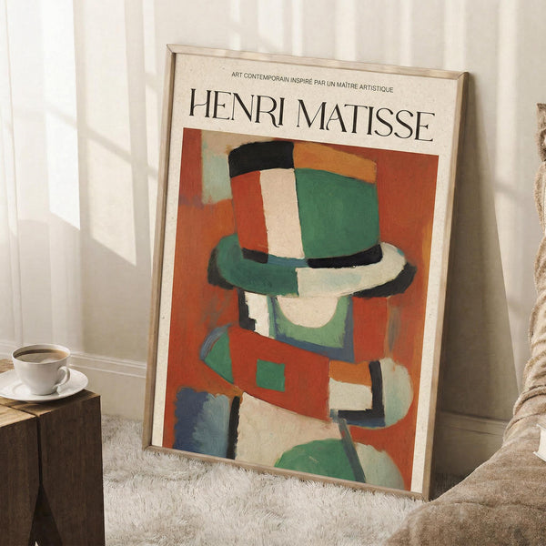 May include: Framed Henri Matisse poster leaning against white wall corner on textured white rug. Abstract artwork shows colorful geometric shapes in green, orange, red, and cream forming a stylized figure wearing a hat. Natural wood frame surrounds the vintage-style art print. Small wooden side table with white coffee cup and saucer sits nearby, with cozy beige throw blanket visible at edge.