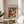 May include: Framed Henri Matisse poster featuring abstract colorful artwork with geometric shapes in green, red, orange, and blue tones, displayed on a light wood round side table next to a beige upholstered chair. Natural sunlight streams through white curtains creating soft shadows, with a plant visible in the corner of this minimalist living space.