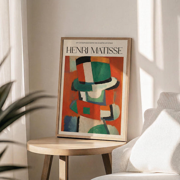 May include: Framed Henri Matisse poster featuring abstract colorful artwork with geometric shapes in green, red, orange, and blue tones, displayed on a light wood round side table next to a beige upholstered chair. Natural sunlight streams through white curtains creating soft shadows, with a plant visible in the corner of this minimalist living space.