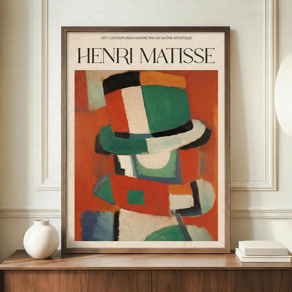 May include: Framed Henri Matisse exhibition poster featuring an abstract figure in colorful geometric shapes - orange, green, red, white, and black patches forming a stylized person wearing a hat. The poster has cream background with black text at top. Displayed on a wooden sideboard next to white ceramic vase and stacked books against cream wall with white molding.