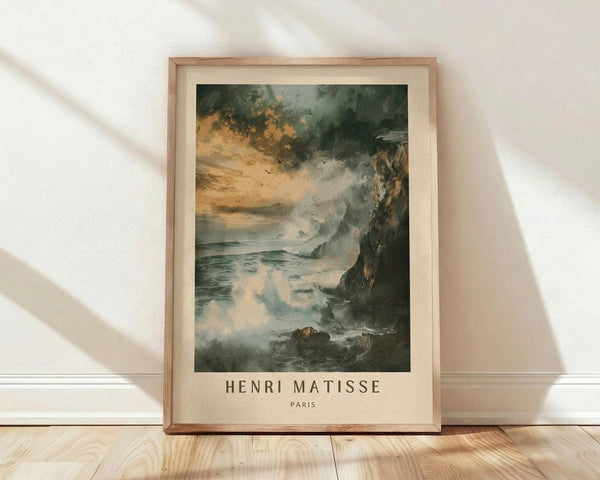 May include: Framed art print showing a dramatic seascape with turbulent storm clouds in green and orange hues above crashing waves against dark rocky cliffs. Small birds fly through the stormy sky. The artwork is displayed in a light wood frame with cream matting, leaning against a white wall on blonde hardwood floors with soft natural lighting casting diagonal shadows.