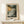 May include: Framed art print showing a dramatic coastal seascape with turbulent waves crashing against dark rocky cliffs under stormy clouds in muted green, gray and cream tones. The poster features "HENRI MATISSE" text below the artwork and sits in a light wood frame on a white wooden surface next to a small white gift box with twine and greenery sprig against a cream wall.