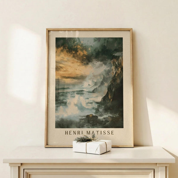 May include: Framed art print showing a dramatic coastal seascape with turbulent waves crashing against dark rocky cliffs under stormy clouds in muted green, gray and cream tones. The poster features "HENRI MATISSE" text below the artwork and sits in a light wood frame on a white wooden surface next to a small white gift box with twine and greenery sprig against a cream wall.