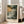 May include: Framed art print showing a dramatic coastal landscape with rocky cliffs, turbulent ocean waves, and stormy sky in muted greens, grays, and warm golden tones. The poster features "HENRI MATISSE PARIS" text below the artwork and sits in a light wooden frame against a beige sofa in a bright, minimalist living room with white walls and large windows casting natural light.