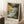 May include: Framed poster print of a dramatic seascape with turbulent waves crashing against dark cliffs under stormy golden and green clouds with flying birds, displayed leaning against white wall next to coffee cup on wooden block and cream blanket on white textured rug