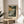 May include: Framed art print featuring a moody seascape with dramatic green and amber clouds above rocky coastal cliffs and turbulent waters, displayed on a round wooden side table next to a beige upholstered chair in a sunlit room with white walls and plant shadows.