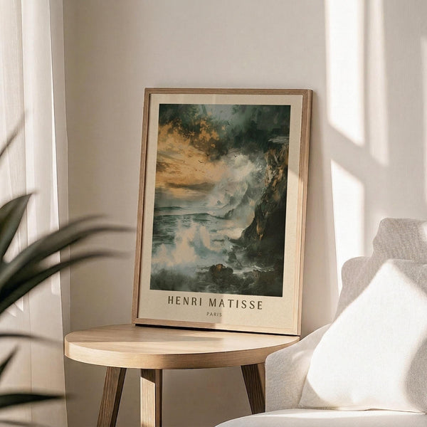 May include: Framed art print featuring a moody seascape with dramatic green and amber clouds above rocky coastal cliffs and turbulent waters, displayed on a round wooden side table next to a beige upholstered chair in a sunlit room with white walls and plant shadows.