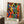 May include: Framed poster with vibrant abstract figures in silhouette dancing in blues, greens, oranges, and yellows, featuring "MATISSE" text at bottom in green letters. Light wooden frame leaning against white wall corner next to beige armchair and small wooden side table with white coffee cup and books.