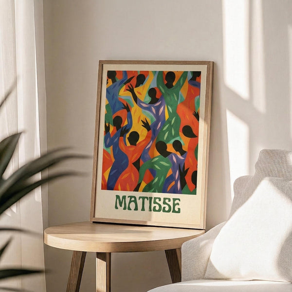 May include: Colorful art print in wooden frame displaying abstract dancing figures in vibrant blues, greens, oranges, and yellows with black silhouettes, artist name below artwork. Frame sits on round wooden side table next to beige upholstered chair with plant shadows cast on white wall behind.