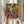 May include: Hands holding a framed colorful poster featuring dancing figures in silhouette with vibrant blues, greens, oranges, and reds in an abstract, flowing style. The artwork shows black silhouetted people with raised arms against bright geometric backgrounds. Green "MATISSE" text appears at bottom. Person wears cream ruffled sleeves and holds dried wheat stems. White curtained window visible in background.