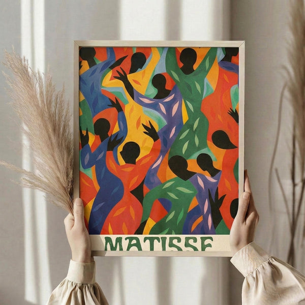 May include: Hands holding a framed colorful poster featuring dancing figures in silhouette with vibrant blues, greens, oranges, and reds in an abstract, flowing style. The artwork shows black silhouetted people with raised arms against bright geometric backgrounds. Green "MATISSE" text appears at bottom. Person wears cream ruffled sleeves and holds dried wheat stems. White curtained window visible in background.