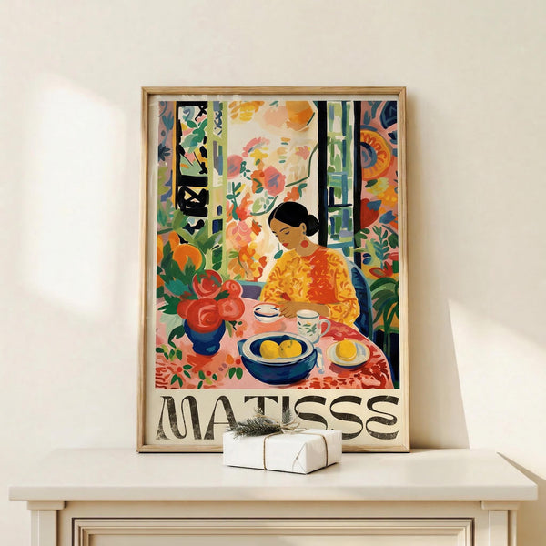 May include: Framed poster featuring a colorful Fauve-style interior scene with a woman in yellow seated at a table with fruit bowl, teacup, and red flowers in vase. Vibrant oranges, greens, and blues fill the room with decorative patterns. Natural wood frame sits on white shelf beside small wrapped gift with sprig decoration against cream wall.