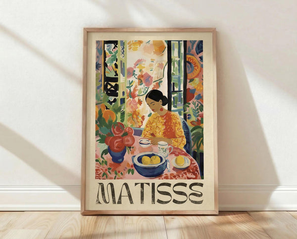 May include: Framed art print showing a colorful interior scene with a woman in yellow dress sitting at a table with fruit bowl, teacup, and red flowers in blue vase. Vibrant Post-Impressionist style with bold colors, patterns, and foliage visible through windows. Text reads "MATISSE" below image. Light wood frame against white wall on wooden floor with soft natural lighting.